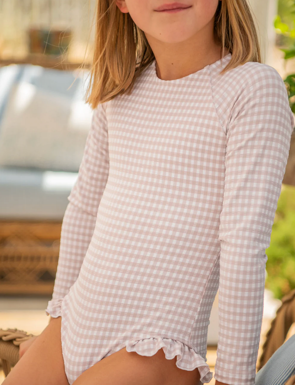 A child in a long-sleeve Minnow Girls' Rashguard One Piece swimsuit with pink checkered pattern, grosgrain ribbon accents, and ruffled edges sits outdoors, enjoying the sun while staying sun-safe.