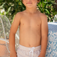 A young boy wearing the Minnow Boy's Boardie stands next to a table with a patterned cloth, surrounded by lush greenery, perfectly embodying style while enjoying unparalleled comfort and mobility.