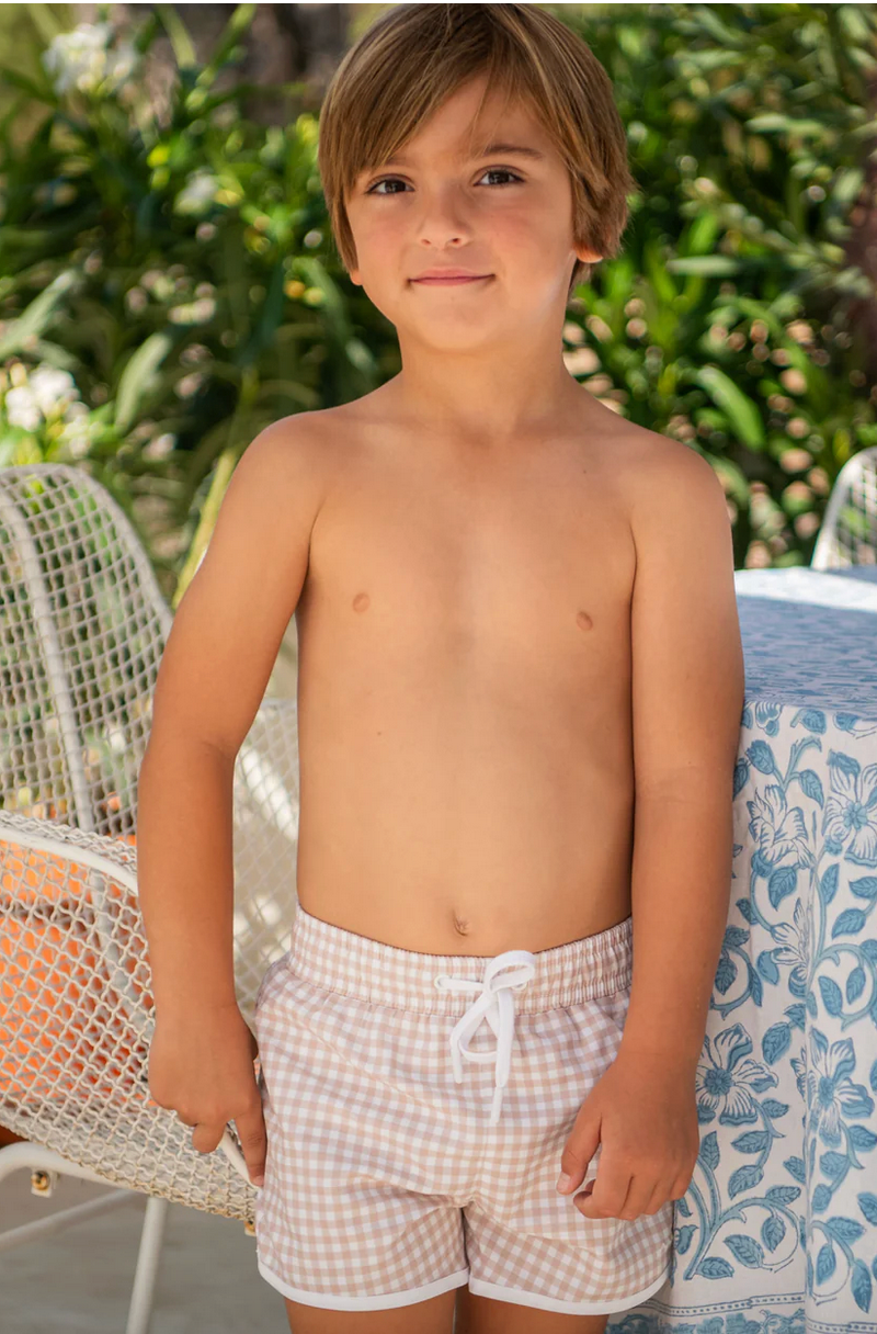 A young boy wearing the Minnow Boy's Boardie stands next to a table with a patterned cloth, surrounded by lush greenery, perfectly embodying style while enjoying unparalleled comfort and mobility.