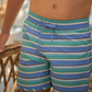 A young boy, embodying a laid-back style, stands next to a wicker chair in his Minnow Boy's Boardie swim trunks featuring blue and green stripes. He's ready for the sun with his UPF 50+ rashguard from Minnow, ensuring comfort and mobility for whatever seaside adventures await.