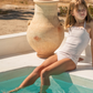 A girl in the Minnow Girls Halter One Piece with Tie Knot sits by the pool, dipping her legs in the water next to a large ceramic vase, basking under the warm sunlight.