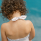 A curly-haired child in a checkered Minnow Girls Halter One Piece with Tie Knot stands near a body of water.