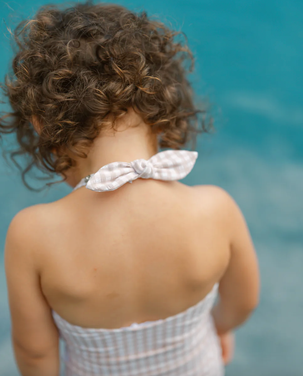 A curly-haired child in a checkered Minnow Girls Halter One Piece with Tie Knot stands near a body of water.