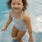 A child wearing a Minnow Girls Halter One Piece with Tie Knot swimsuit from the brand Minnow stands in shallow water, smiling at the camera.