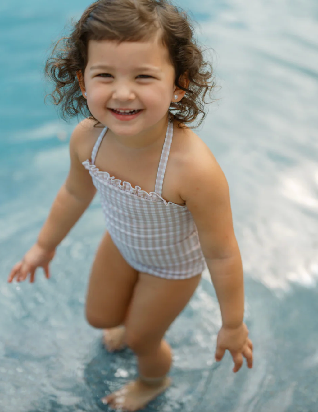 A child wearing a Minnow Girls Halter One Piece with Tie Knot swimsuit from the brand Minnow stands in shallow water, smiling at the camera.