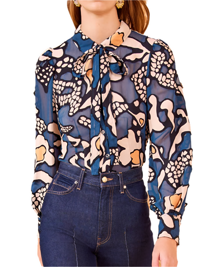 A person wearing the Ulla Johnson Hera Blouse featuring floral motifs and a bow tie, paired with high-waisted jeans, against a plain background.