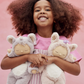 A child with curly hair smiles joyfully, holding two Olli Ella Cozy Dinkum Unicorn plush toys with irresistibly soft fur, ready for magical adventures against a pink background.