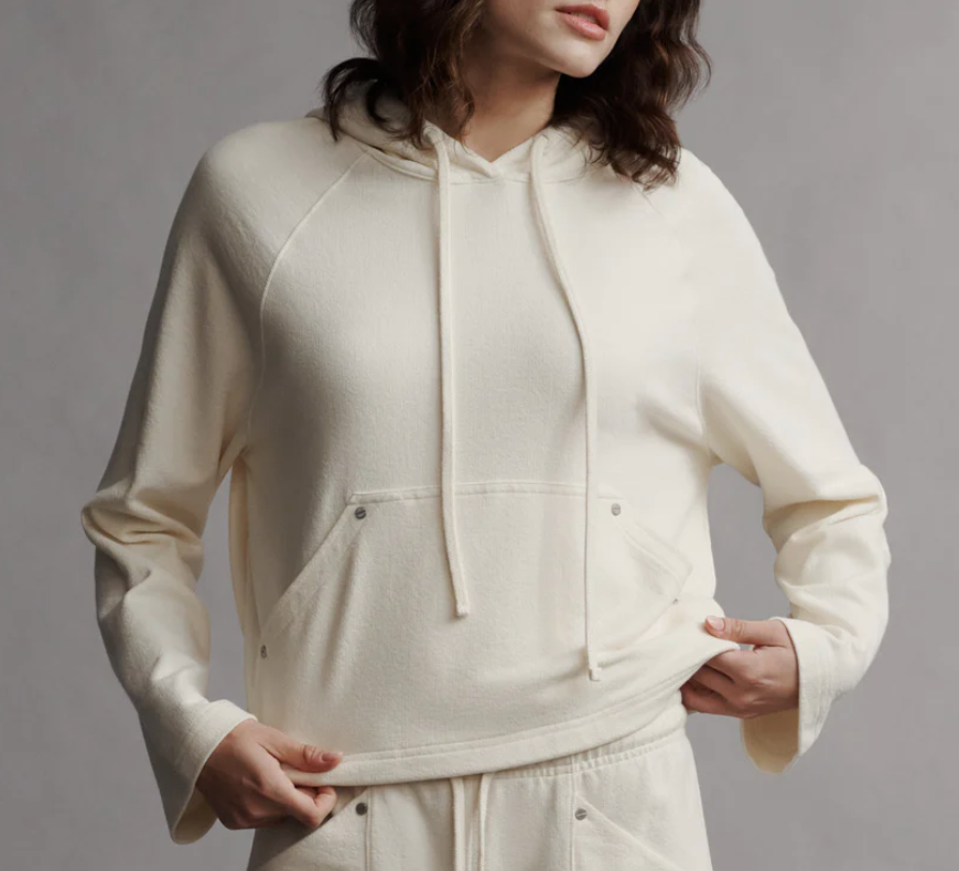 A person is wearing TWP Ali's Hoodie, featuring a relaxed cream color and slouchy fit with hands tucked in the pockets. The French Terry fabric enhances comfort, while the drawstring adjustable hood completes the casual look.