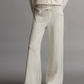 A person wearing the TWP Saturday Night Pant, featuring a mid-rise, loose fit in beige with an elasticated waistband and hands in pockets, stands against a plain gray background.