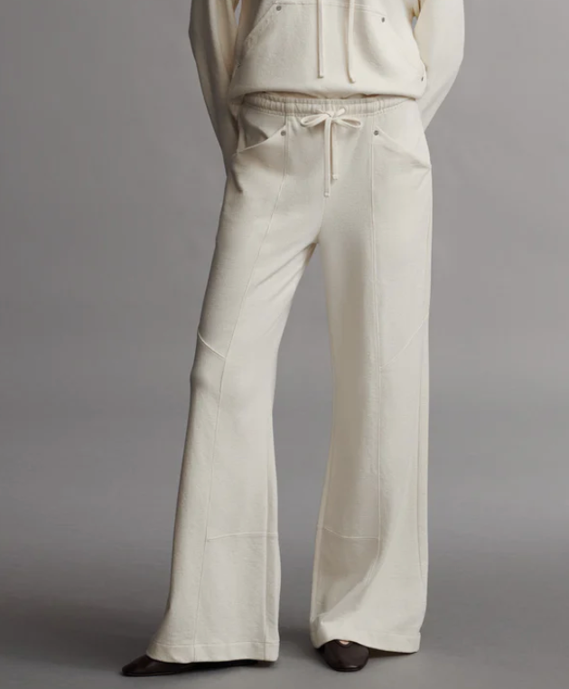 A person wearing the TWP Saturday Night Pant, featuring a mid-rise, loose fit in beige with an elasticated waistband and hands in pockets, stands against a plain gray background.