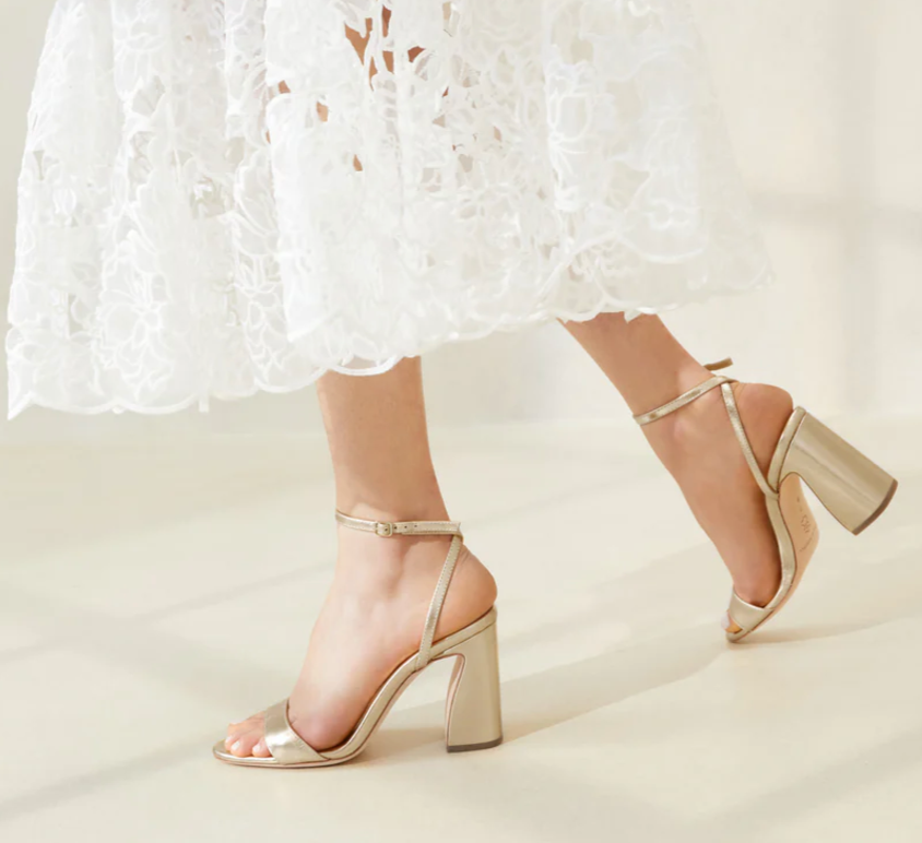 A person in a white lace dress walks indoors on a light-colored floor wearing Loeffler Randall's Malia Curved Heel Sandals, featuring an adjustable ankle strap.