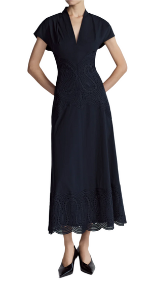 A person in the Lela Rose Poplin V-Neck Dress, featuring an ankle-length design with intricate lace paneled details and an A-line silhouette, stands with hands in pockets while wearing black pointed-toe shoes.