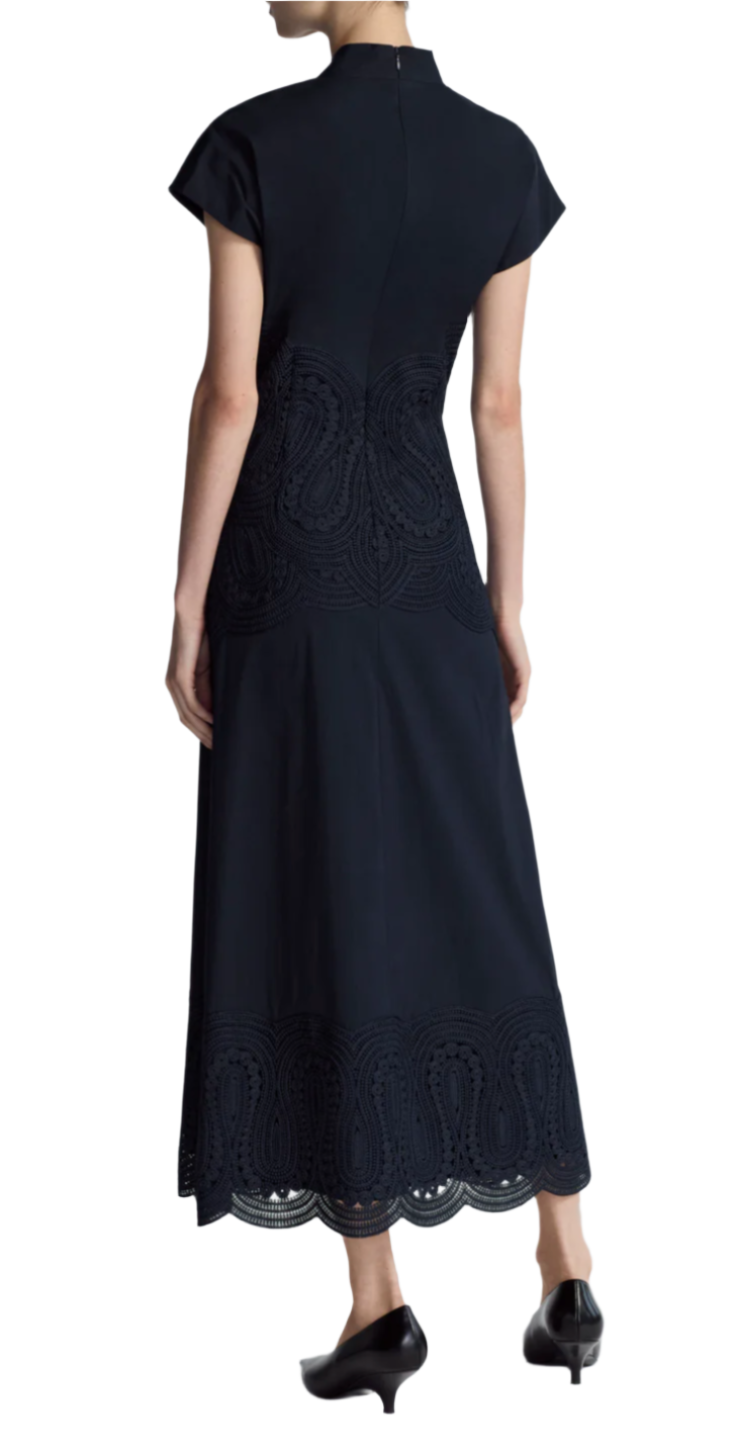 A person is seen from the back, wearing the Lela Rose Poplin V-Neck Dress. This long, dark blue dress features elegant lace-paneled details and an A-line silhouette that provides a graceful flair, complemented by black shoes.