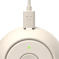 A close-up of the Rest Go Sounds Machine by Hatch, a white, round portable device with a charging cable plugged in and a small green indicator light.