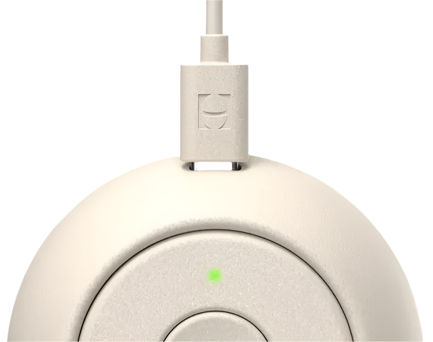 A close-up of the Rest Go Sounds Machine by Hatch, a white, round portable device with a charging cable plugged in and a small green indicator light.