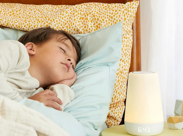 A child sleeps peacefully on light blue sheets in light pajamas. Beside the slumbering youngster, a digital clock reads 6:43, gently illuminated by a soft-glow lamp. Nearby, Hatch's Rest+ 2nd Gen Night Light and Sound Machine provides soothing sounds for an uninterrupted night's rest.