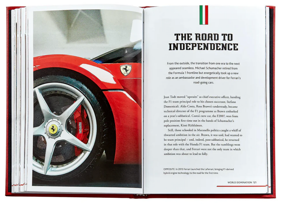 An open book features the wheel of a red sports car on the left page and text about Ferrari's motorsport history on the right, titled "The Road to Independence." Product: The Story Of Ferrari Red Leather by Graphic Image.