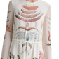 A person wears the Altuzarra Sybbeline Dress, a white silk maxi dress featuring colorful illustrations and French text. The hem is adorned with floral and shell motifs, complemented by a waist tie. Only part of their face is visible.