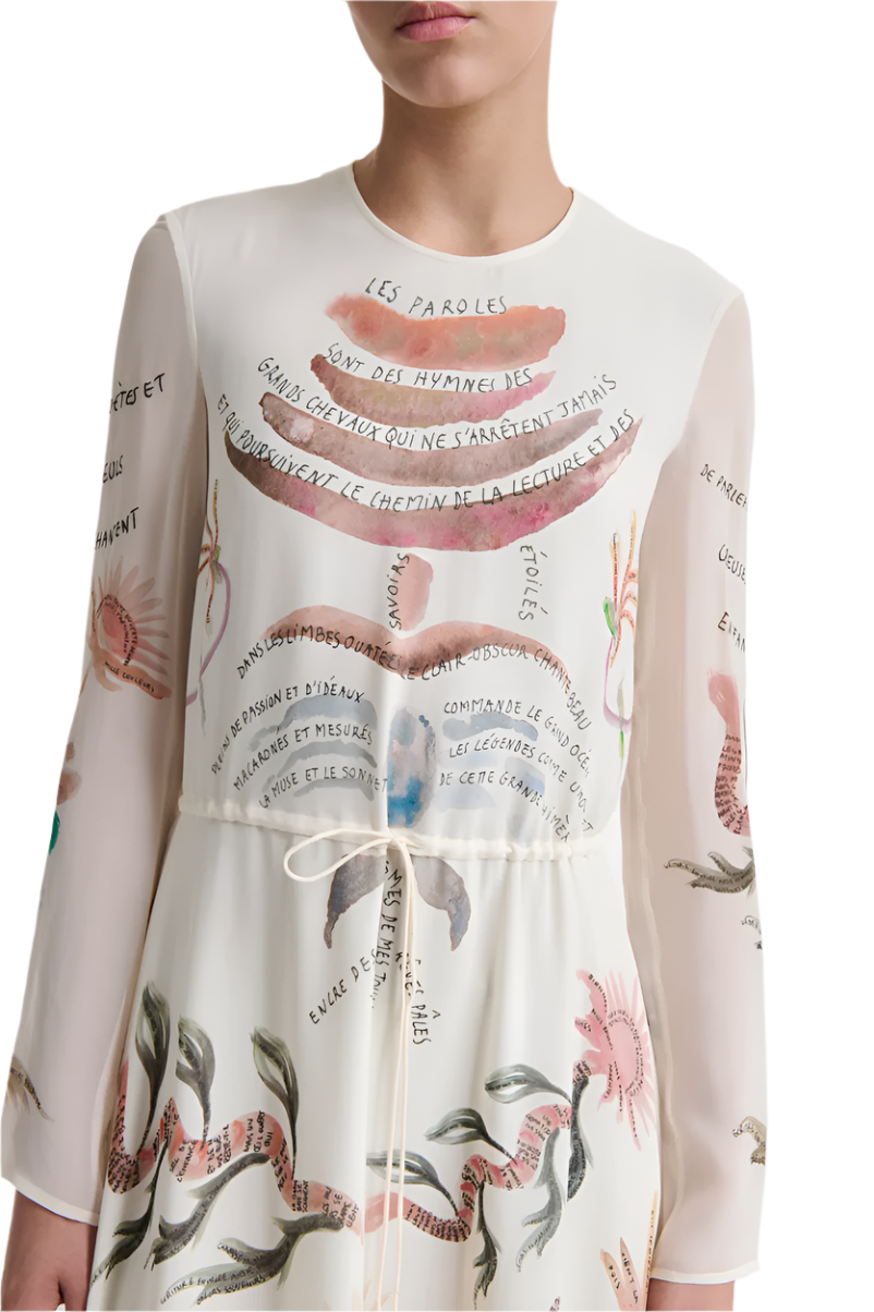 A person wears the Altuzarra Sybbeline Dress, a white silk maxi dress featuring colorful illustrations and French text. The hem is adorned with floral and shell motifs, complemented by a waist tie. Only part of their face is visible.