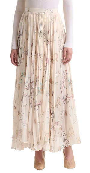 The individual is wearing a white long-sleeve top paired with Altuzarra's high-waisted, pleated Sif Skirt in cream adorned with a floral pattern, elegantly completed with beige shoes.