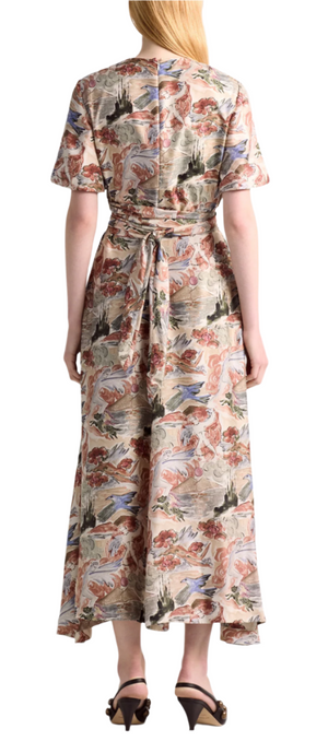 A woman faces away, wearing the Altuzarra Honey Dress—a mid-length, short-sleeve silk dress with a multicolored nature-inspired print—and black heeled sandals with an oversized buckle.