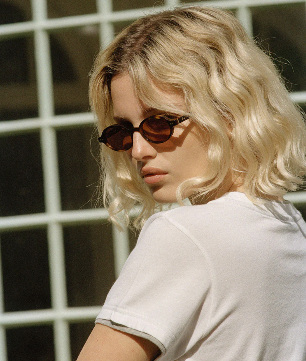 A person with short, wavy blonde hair wears Jimmy Fairly's Elvie Sunglasses—oval-frame shades with metal hinges—and a white T-shirt, standing in front of a window with a grid pattern.