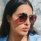 A woman with long dark hair wears Jimmy Fairly Angie Sunglasses, featuring a hexagonal tortoiseshell frame and tinted lenses, paired with a light patterned shirt as she faces sunlight.