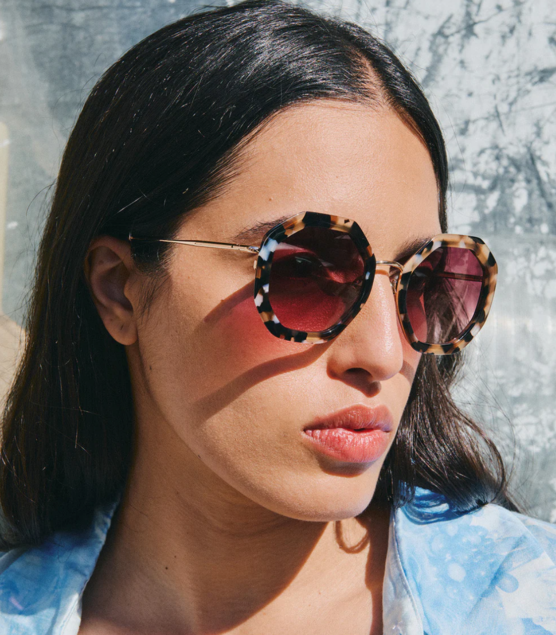 A woman with long dark hair wears Jimmy Fairly Angie Sunglasses, featuring a hexagonal tortoiseshell frame and tinted lenses, paired with a light patterned shirt as she faces sunlight.