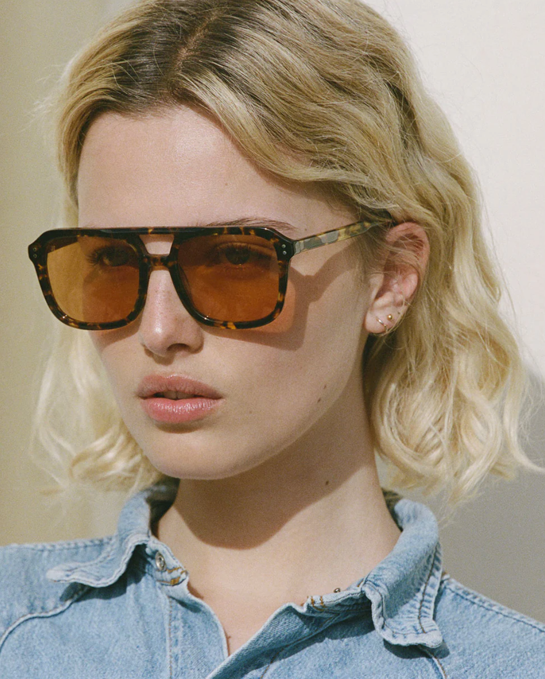 A person with short blonde hair wears Jimmy Fairly Elma JF Colors sunglasses with large brown-tinted cellulose acetate Aviator frames and a light blue denim shirt, looking ahead with a neutral expression.