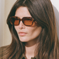 Woman with long brown hair wears Jimmy Fairly Freja Sunglasses in brown cellulose acetate and a brown collared shirt, gazing slightly to the side in soft lighting.