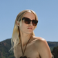 A woman with long blonde hair wears Jimmy Fairly’s Maya Sunglasses with a chic butterfly frame, looking to the side outdoors against a clear blue sky and green hills.