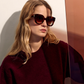 A person with long, wavy blonde hair wears Jimmy Fairly's Swirl Sunglasses with a butterfly frame and a burgundy sweater while sitting next to a reflective, tinted glass panel.