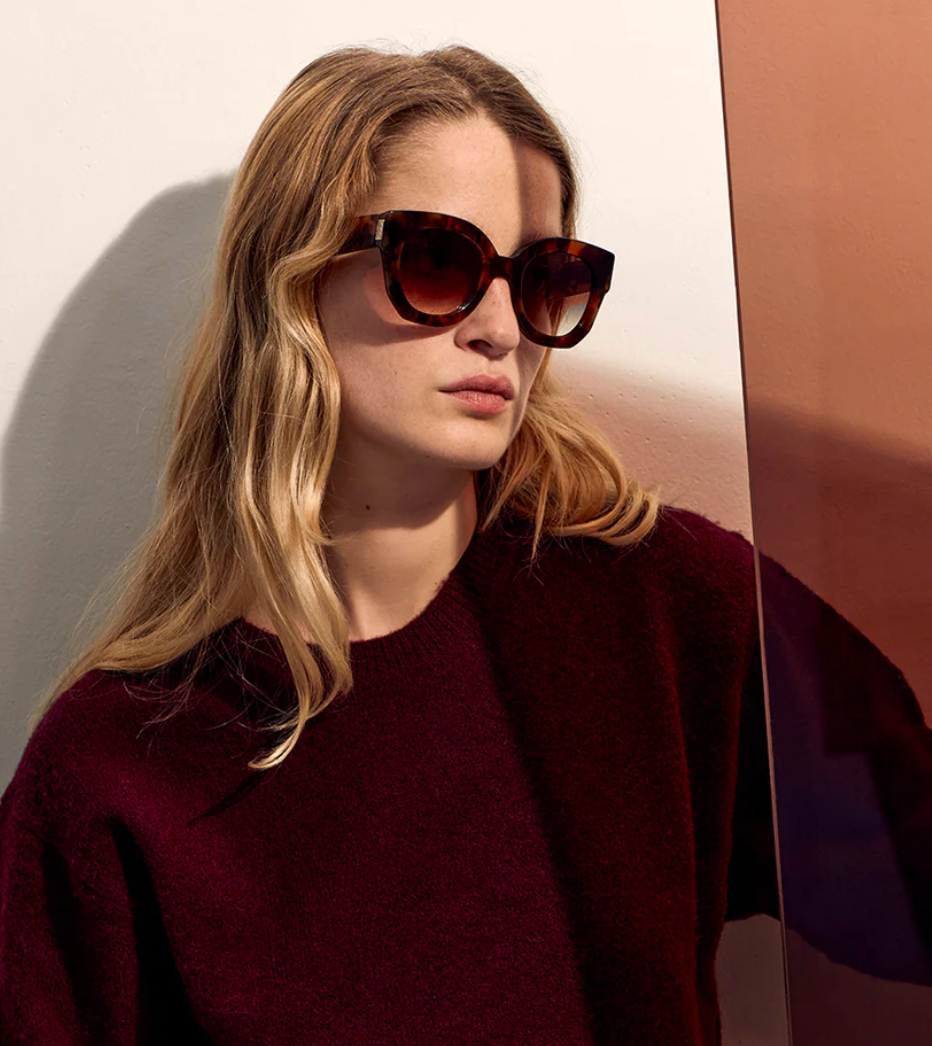 A person with long, wavy blonde hair wears Jimmy Fairly's Swirl Sunglasses with a butterfly frame and a burgundy sweater while sitting next to a reflective, tinted glass panel.
