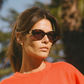 A woman stands outdoors in Jimmy Fairly's Meli Icons Sunglasses, featuring bold butterfly frames and dark lenses, paired with a red top and blurred trees in the background.