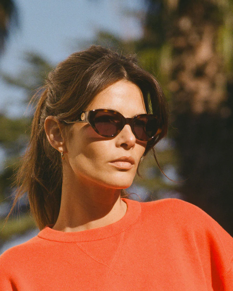 A woman stands outdoors in Jimmy Fairly's Meli Icons Sunglasses, featuring bold butterfly frames and dark lenses, paired with a red top and blurred trees in the background.