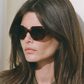 A woman with long brown hair and a dark collared shirt wears Jimmy Fairly's Portofino Sunglasses, featuring hexagonal frames, while standing against a light-colored wall.