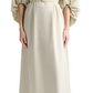 A woman wears the Altuzarra Corrie Dress, featuring voluminous sleeves, a deep V-neckline, a belted waist, and floor-length design, paired with nude flats.