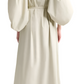 A woman is seen from the back wearing the Altuzarra Corrie Dress, a light-colored maxi with voluminous sleeves and a belt, paired with nude low-heeled shoes.