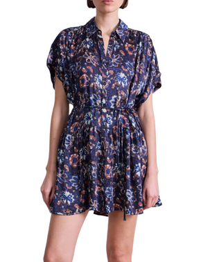 A woman wears the Agnezca Mini Dress by Apiece Apart—a navy blue summer floral print dress with a collar, button-down front, self-tie belt, short sleeves, and a flowy silhouette—standing against a white background.