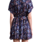 A person is shown from the back wearing the Agnezca Mini Dress by Apiece Apart—a short, loose-fitting, dark blue summer floral dress with a self-tie belt for a cinched waist and flowy silhouette.