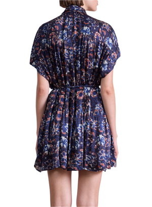 A person is shown from the back wearing the Agnezca Mini Dress by Apiece Apart—a short, loose-fitting, dark blue summer floral dress with a self-tie belt for a cinched waist and flowy silhouette.