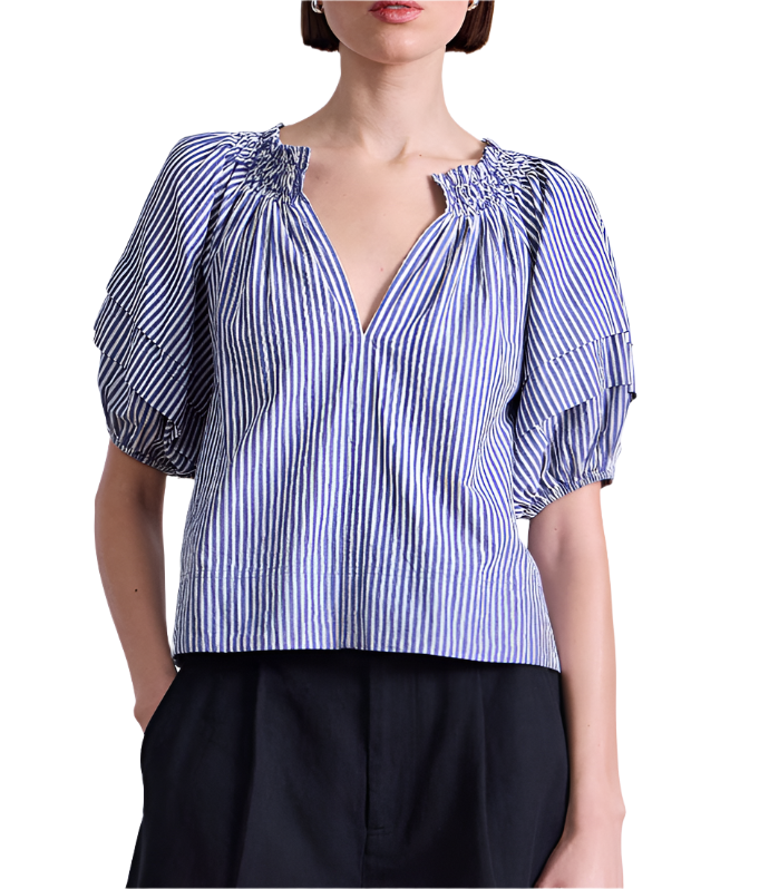 A woman wears the Apiece Apart Caminitas Puff Sleeve Top—a blue and white vertical striped blouse with puffed sleeves and a V-neck—paired with black pants, posing against a white background.