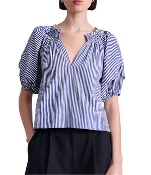 A woman wears the Apiece Apart Caminitas Puff Sleeve Top—a blue and white vertical striped blouse with puffed sleeves and a V-neck—paired with black pants, posing against a white background.