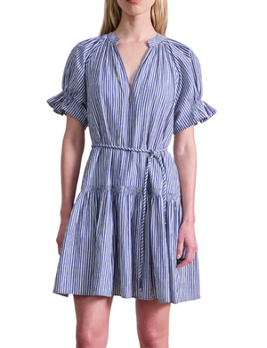 A woman wears the Apiece Apart Marza Mini Dress, featuring blue and white vertical stripes, short ruffled sleeves, a V-neckline, and a matching tie belt at the waist.