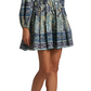 A woman wears the Ulla Johnson Anika Dress—a patchwork print mini dress with blouson sleeves, a V-neck, and a tiered skirt—paired with yellow high-heeled sandals adorned with playful pom-pom accents.