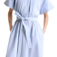 A person wearing the Altuzarra Paulina Banker Stripe Dress—a light blue, short-sleeve cotton poplin shirt dress with thin white vertical stripes, belted waist, and hands in pockets.