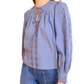 A woman wears the Ulla Johnson Kady Blouse, a light blue, long-sleeve top in water-washed cotton with geometric embroidery, paired with high-waisted blue jeans against a white background.