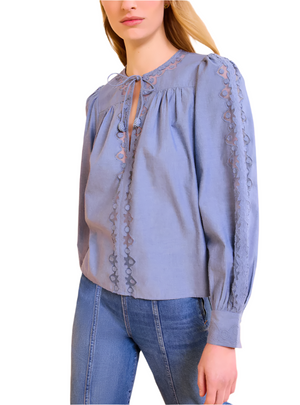A woman wears the Ulla Johnson Kady Blouse, a light blue, long-sleeve top in water-washed cotton with geometric embroidery, paired with high-waisted blue jeans against a white background.