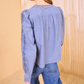 A person stands facing away from the camera, wearing Ulla Johnson's light blue Kady Blouse featuring lace details and geometric embroidery on the sleeves and shoulders, paired with blue jeans.
