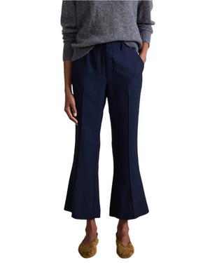 A person models the Apiece Apart Rene Flare Trouser—navy kick flare cropped pants—paired with a grey sweater and brown flats, standing with hands in pockets against a white background.